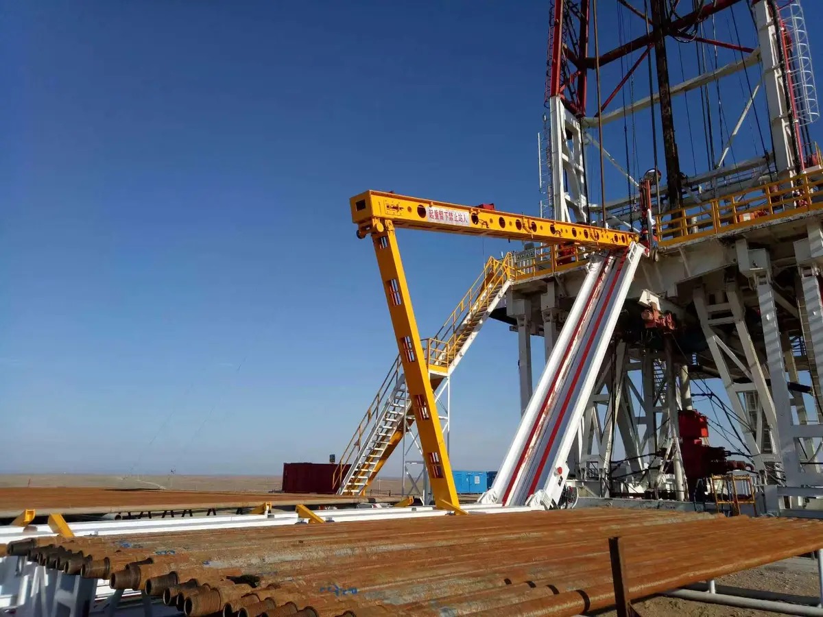 Electric VFD power catwalk raised to the rig floor on a land drilling rig — yellow catwalk delivering drill pipe under blue sky, red derrick in background